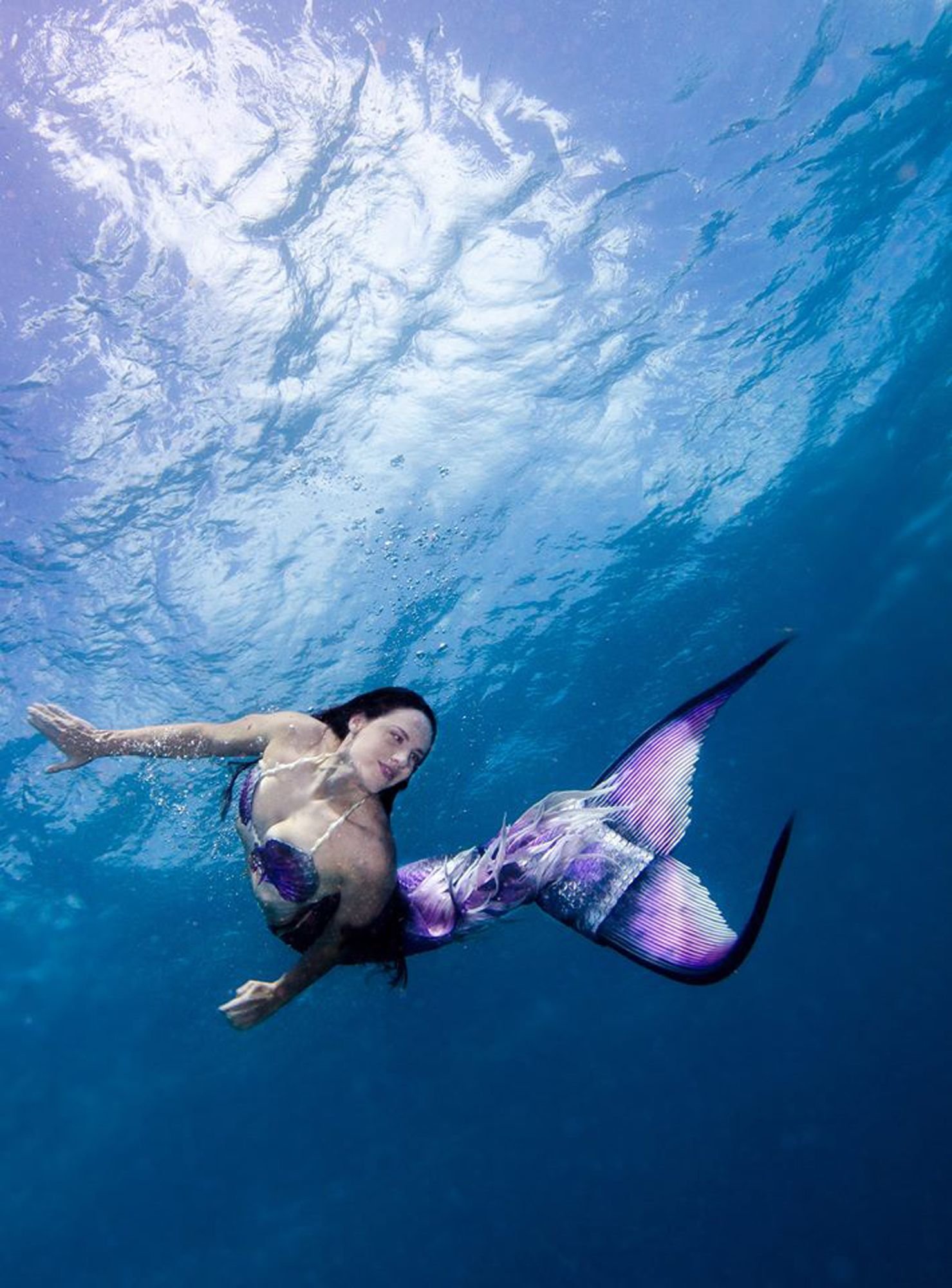 Mermaid model with a purple and silver tail swimming underwater near the surface, light streaming down through the water