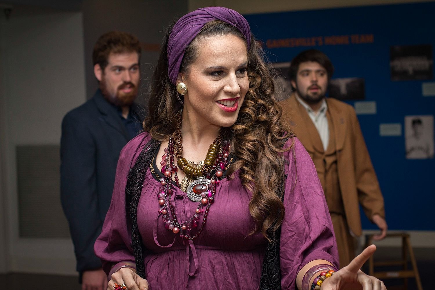Shannon Scott in costume as a bohemian/gypsy-style character with a purple headwrap and layered jewelry, performing on stage with two actors in the background