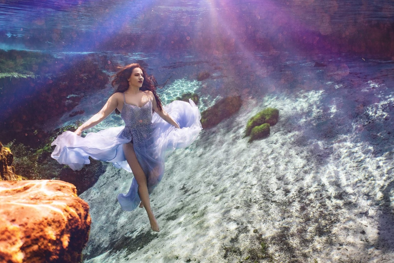Underwater fashion model floating in a flowing light blue gown over a rocky, sunlit spring floor, with hair and fabric drifting in the water