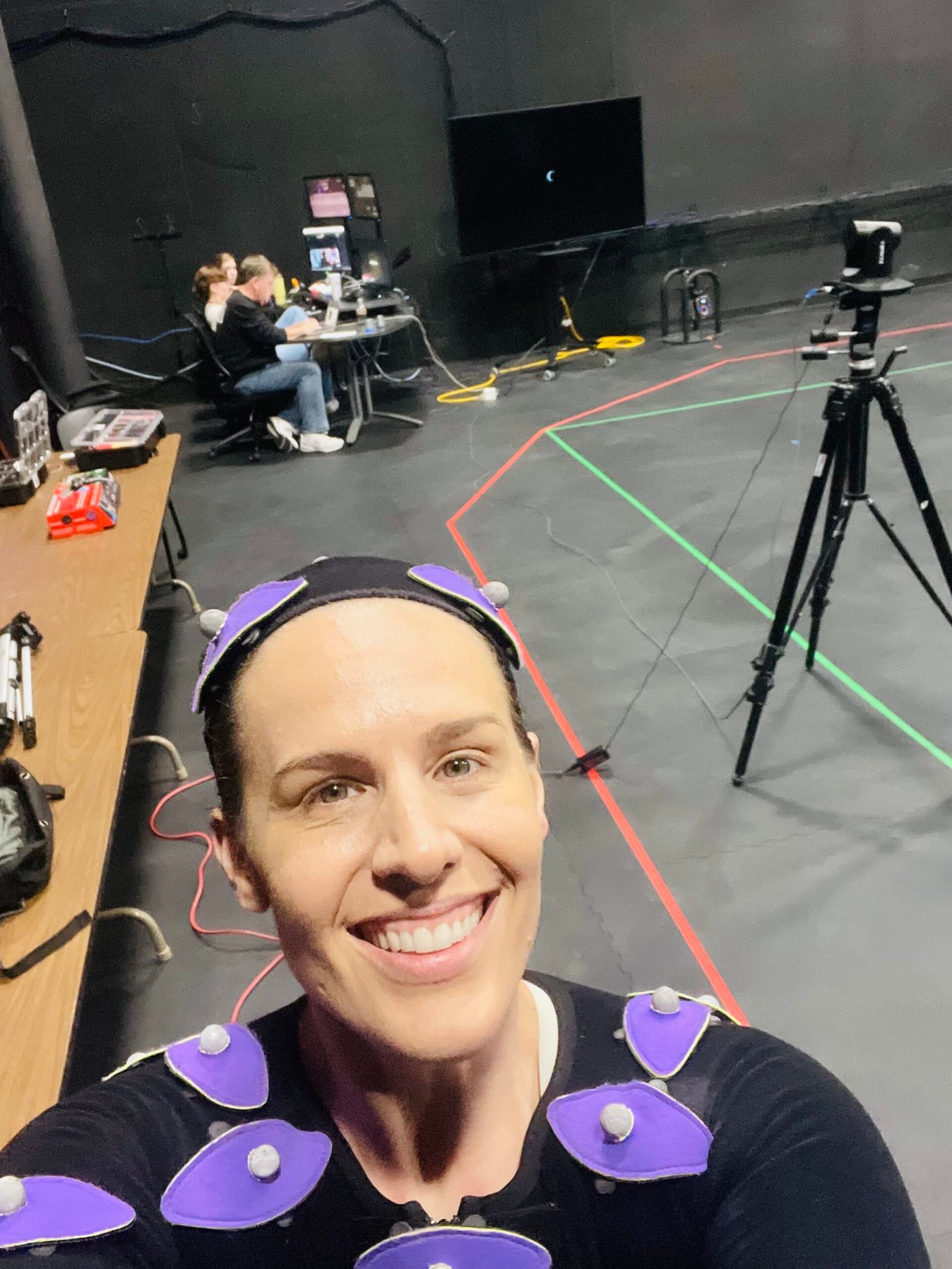 Motion Capture Actress Shannon Scott smiles in her MOCAP suit while standing on a performance capture stage with the production crew preparing the project at computer workstations in the background.